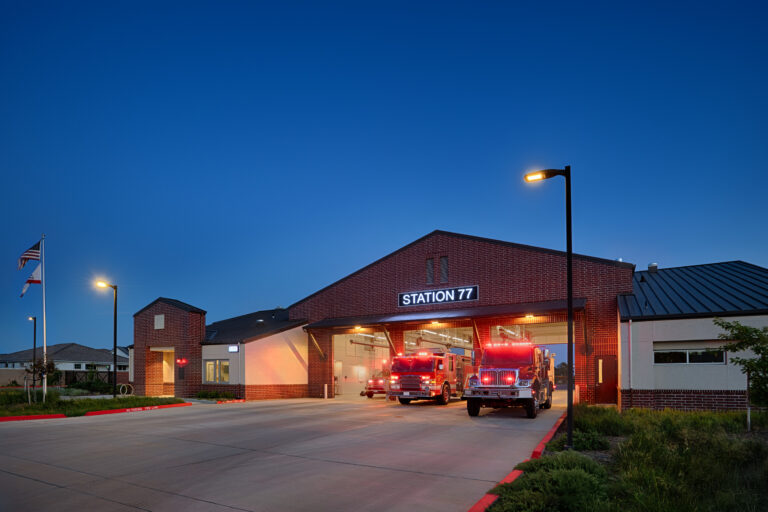 Fire trucks with flashing lights parked outside Station 77 at dusk.