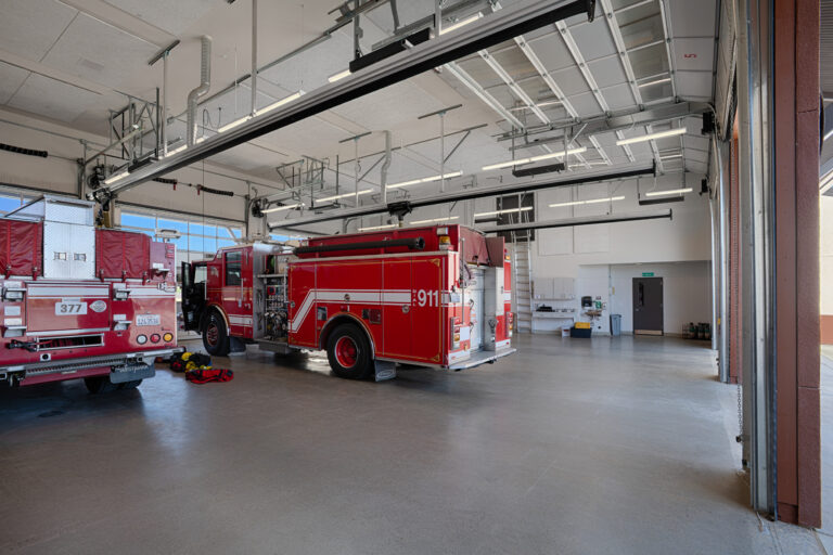 Two red fire engines parked inside a spacious fire station garage with open bay doors and overhead tracks.