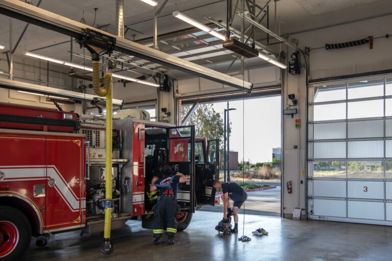 Firefighters gear up beside a red fire engine inside a station with open bay doors and overhead exhaust hoses.