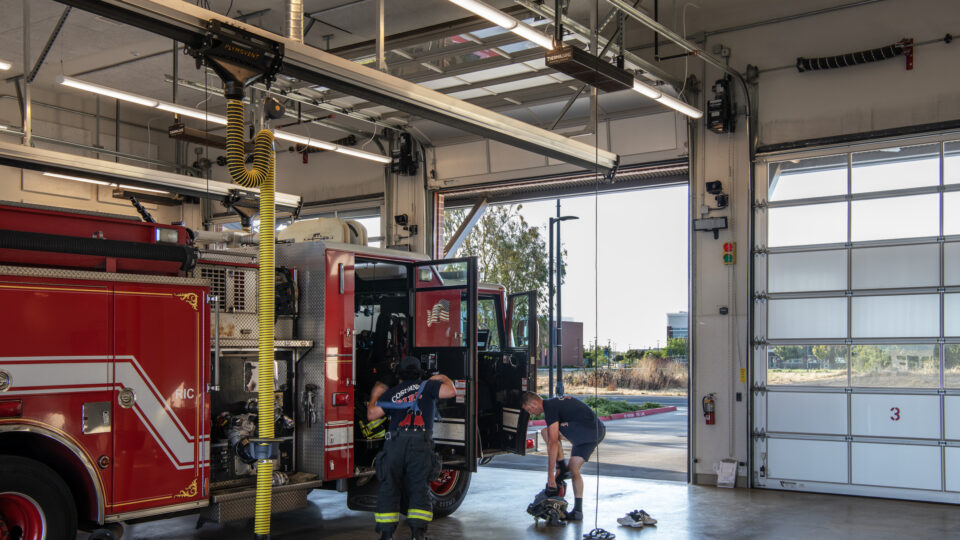 Firefighters gear up beside a red fire engine inside a station with open bay doors and overhead exhaust hoses.