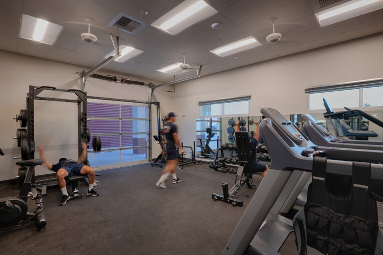 Indoor gym with treadmills, weights, and a bench press, with people exercising under bright ceiling lights.
