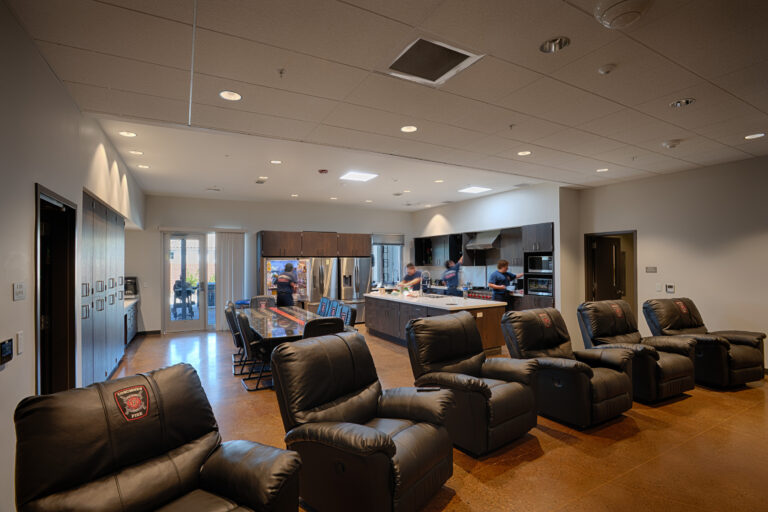 Fire station lounge with black recliners, a dining table, and a modern kitchen where firefighters are cooking