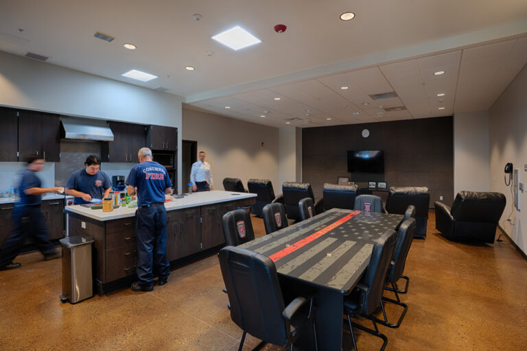 Firefighters preparing food in a modern station kitchen and lounge with a large flag-themed conference table and recliners