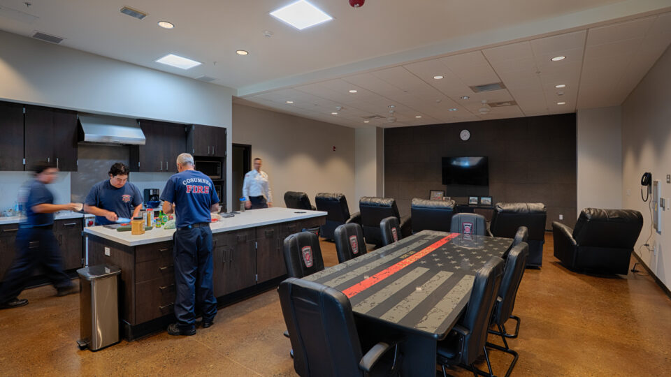 Firefighters preparing food in a modern station kitchen and lounge with a large flag-themed conference table and recliners