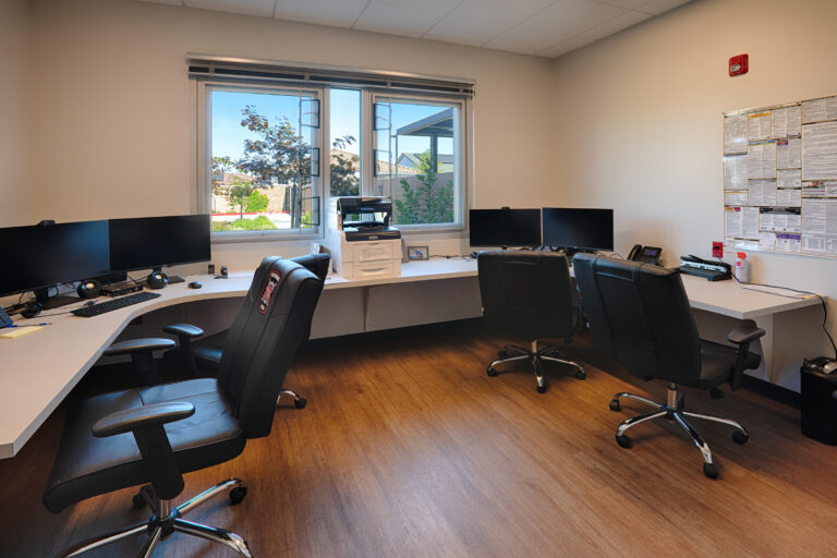 Small office with curved white desks, multiple computer monitors, black chairs, and a large window letting in daylight.