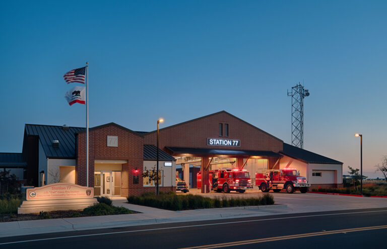 Brick fire station labeled Station 77 at dusk, with two fire engines in the open bay and U.S. and California flags outside