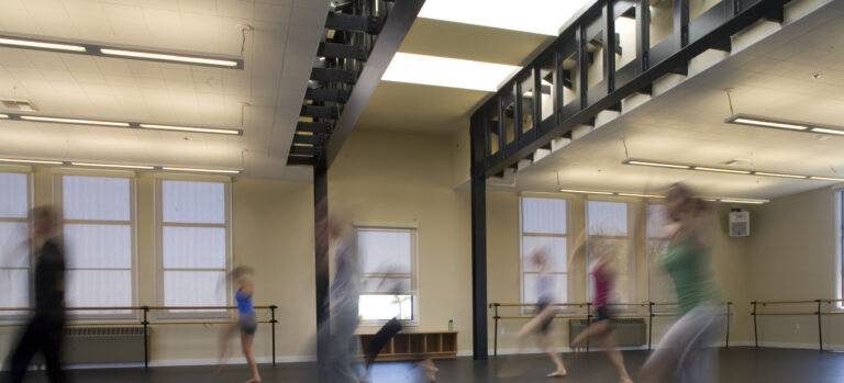 Blurred dancers moving across a spacious studio with ballet barres, tall windows, and an upper walkway