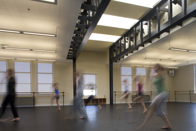Blurred dancers moving across a spacious studio with ballet barres, tall windows, and an upper walkway