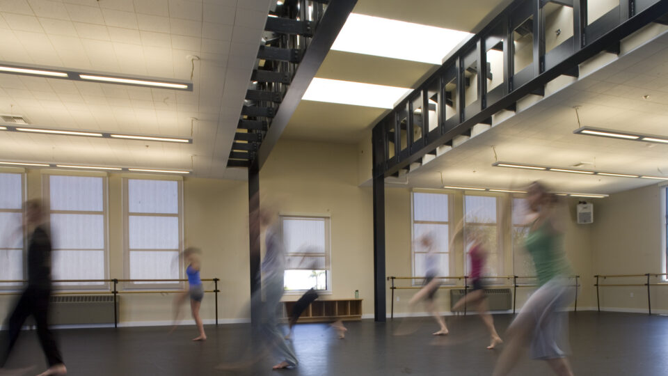 Blurred dancers moving across a spacious studio with ballet barres, tall windows, and an upper walkway