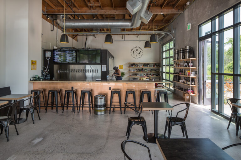 Modern bottle shop and tasting room with a long tap bar, black stools, shelves of bottles, and large windows.