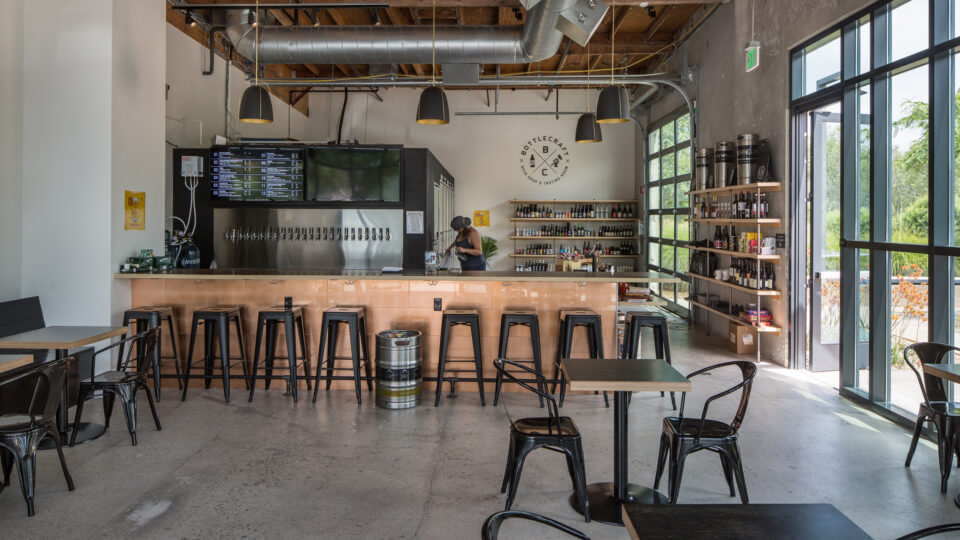 Modern bottle shop and tasting room with a long tap bar, black stools, shelves of bottles, and large windows.