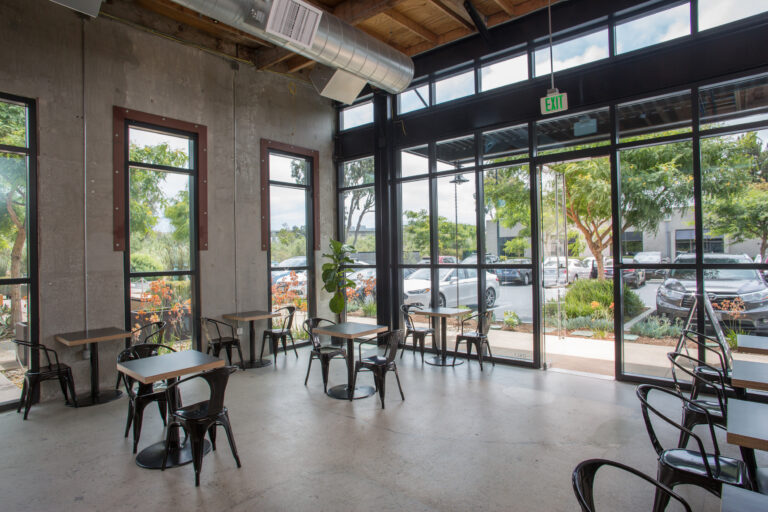 Modern cafe interior with small tables, black metal chairs, concrete walls, and large windows by the entrance.