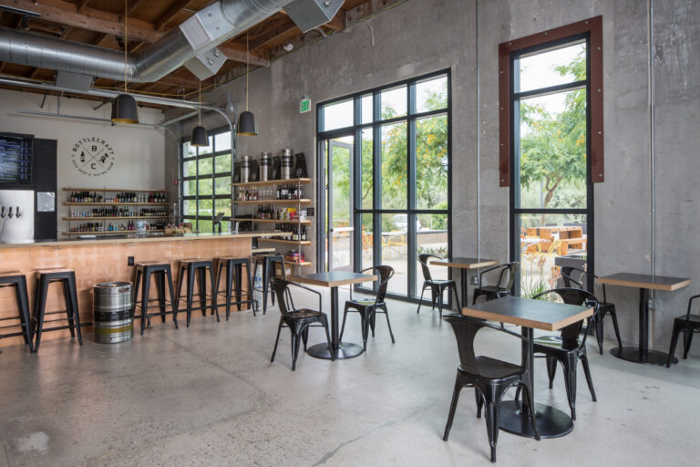 Modern bottle shop and tasting room interior with bar seating, small tables, and large windows overlooking a patio.