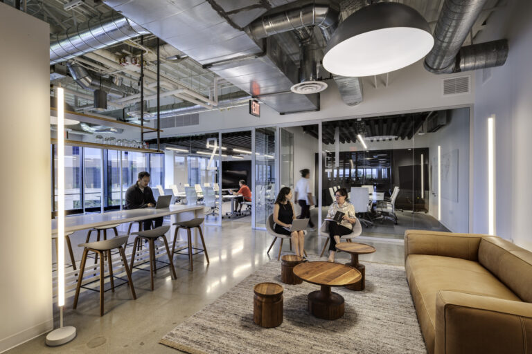 Modern open office lounge with glass meeting rooms, exposed ductwork, high table seating, and people working and talking