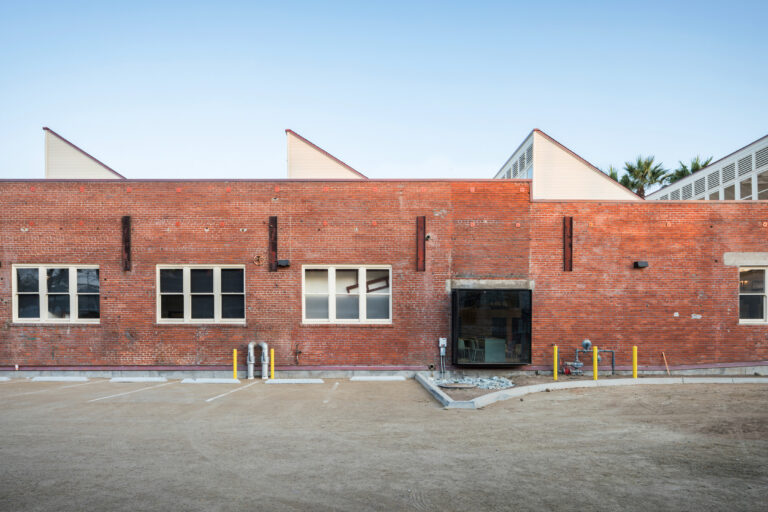 Red brick industrial building with white windows, a glass entrance, and an empty dirt parking lot in front