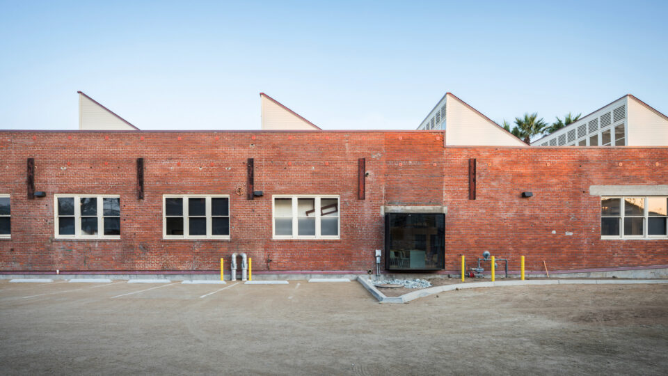 Red brick industrial building with white windows, a glass entrance, and an empty dirt parking lot in front