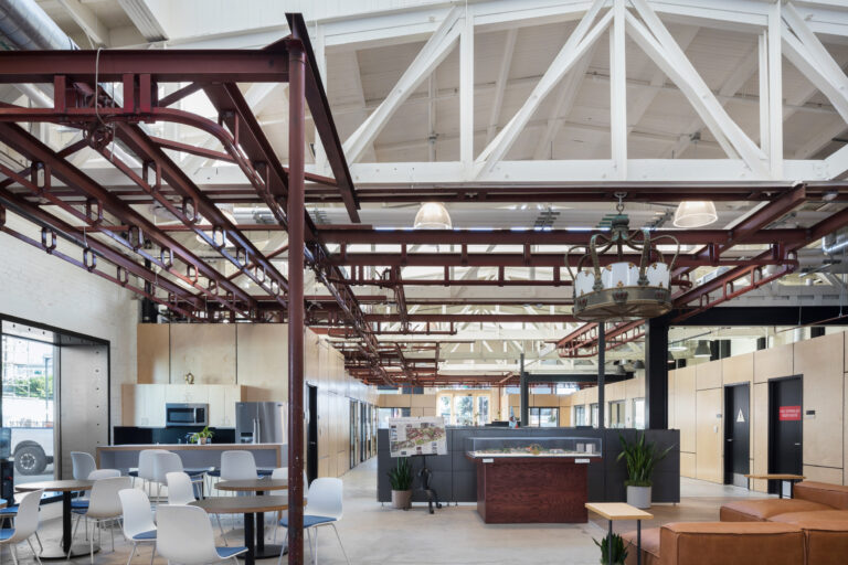 Open industrial-style office atrium with exposed white trusses, red steel beams, café seating, and a leather sofa