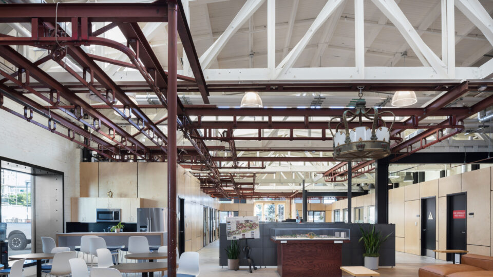 Open industrial-style office atrium with exposed white trusses, red steel beams, café seating, and a leather sofa