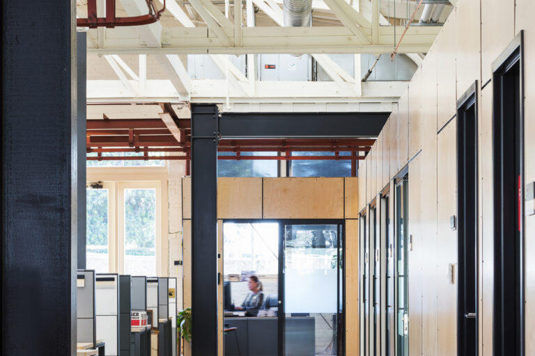 Industrial-style office hallway with exposed beams and ducts, glass-walled rooms, cubicles, and a seated worker at the end
