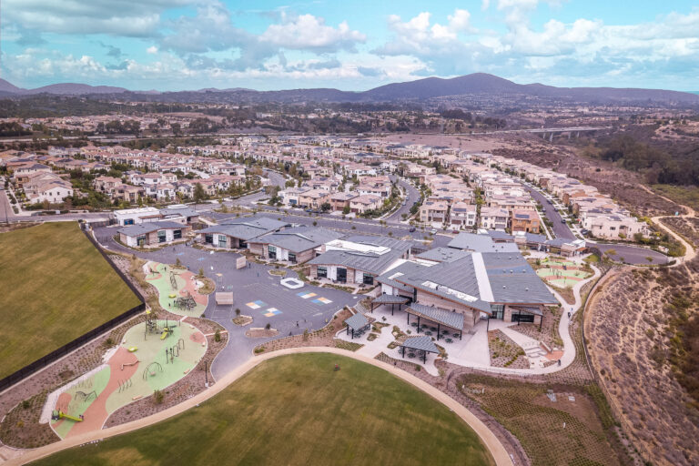 Aerial view of a suburban school campus with playgrounds, sports fields, and surrounding homes against distant hills.