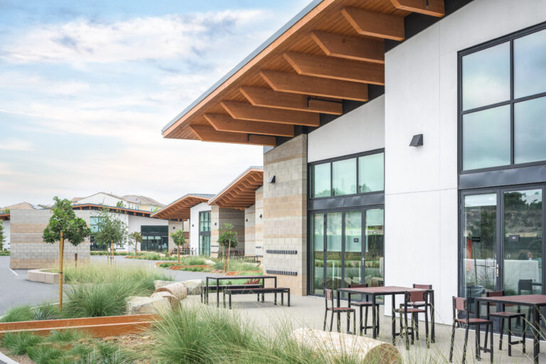 Modern commercial courtyard with white and stone buildings, large windows, wood roof overhangs, and outdoor tables.