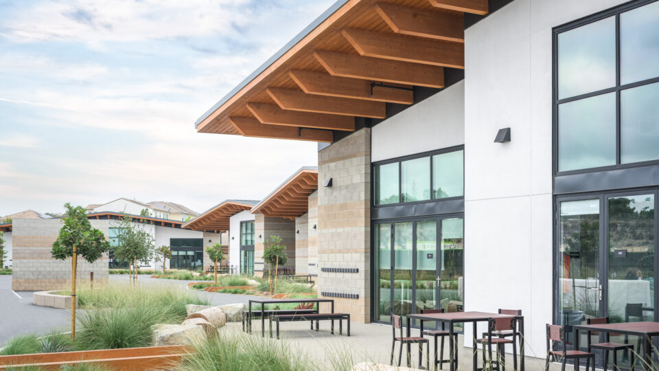 Modern commercial courtyard with white and stone buildings, large windows, wood roof overhangs, and outdoor tables.
