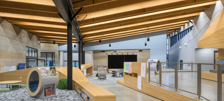 Modern library or school commons with curved wood seating, a small stage, and a high timber beam ceiling