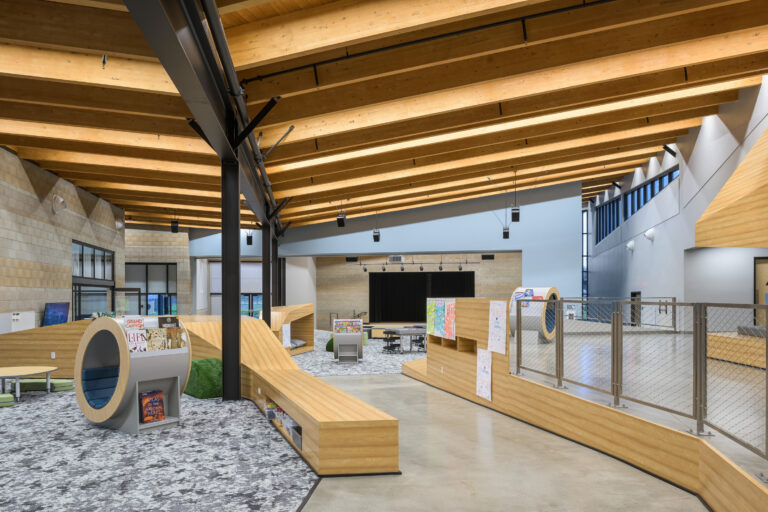 Modern library or school commons with curved wood seating, a small stage, and a high timber beam ceiling