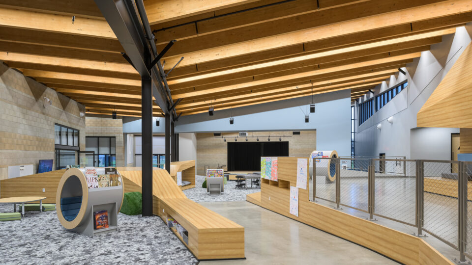 Modern library or school commons with curved wood seating, a small stage, and a high timber beam ceiling