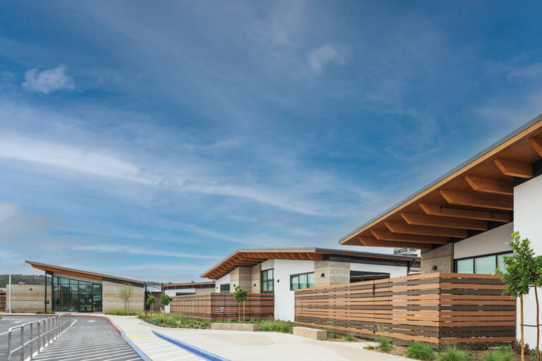 Modern low-rise buildings with wood-accented roofs and fences along a landscaped walkway under a wide blue sky.