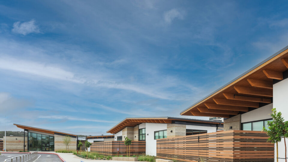 Modern low-rise buildings with wood-accented roofs and fences along a landscaped walkway under a wide blue sky.