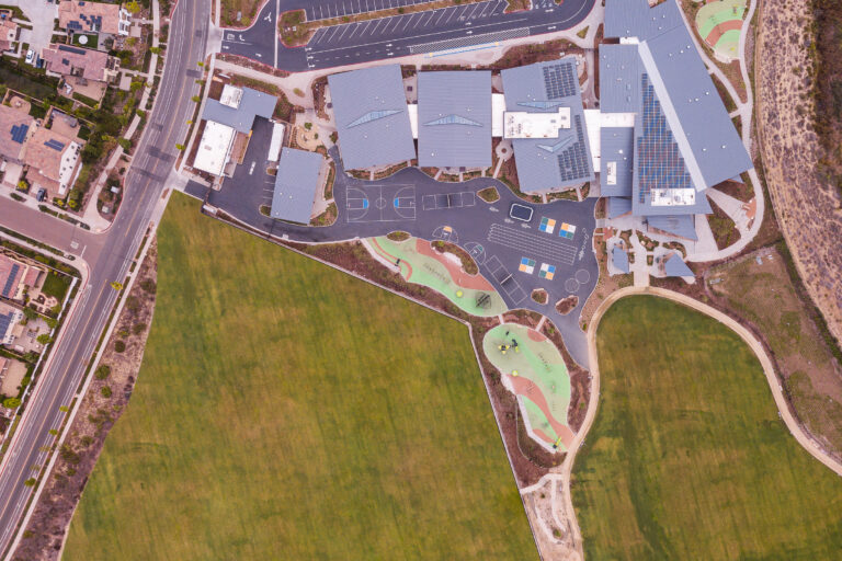 Aerial view of a school campus with buildings, parking lot, play areas, basketball court, and large grassy fields