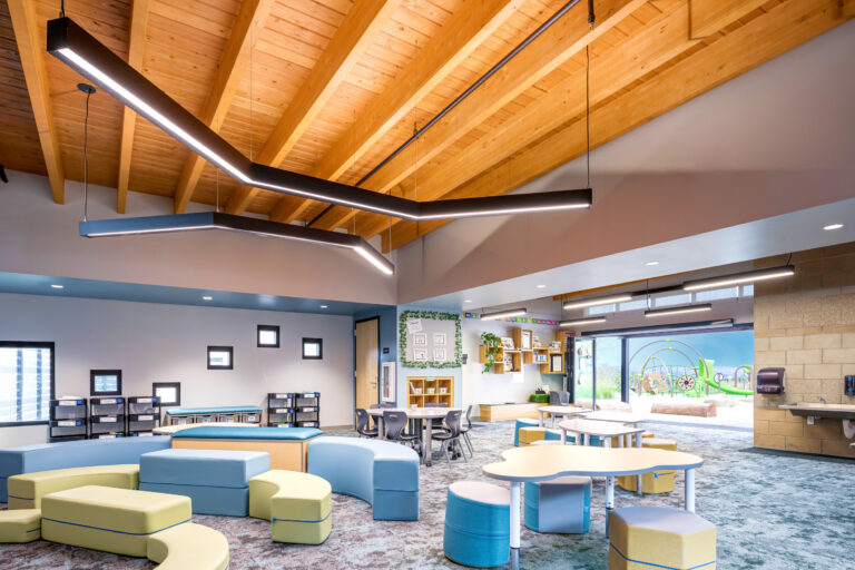 Bright children's learning space with curved seating, small tables, exposed wood ceiling, and playground visible outside.