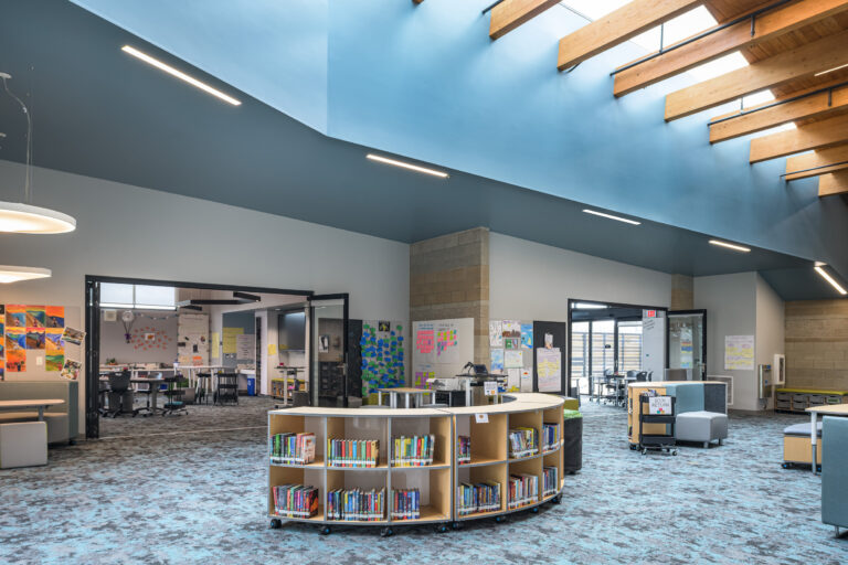 Modern school library with curved low bookshelves, open classrooms, and a high blue ceiling with wood beams and skylights