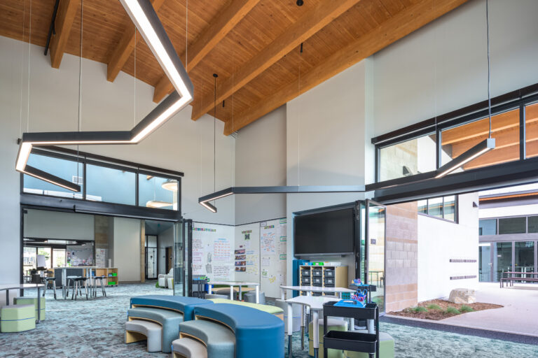 Modern classroom commons with high wood-beam ceiling, geometric pendant lights, flexible seating, and large glass walls.