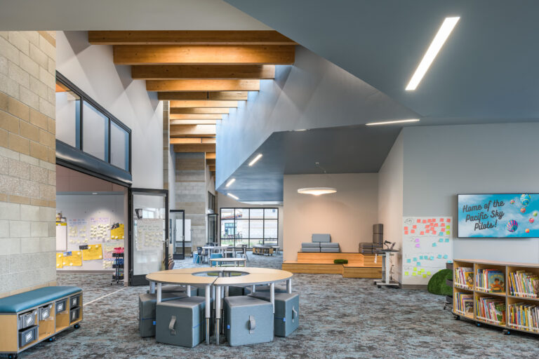 Modern school library interior with round tables, low stools, bookshelves, and a screen reading Home of the Pacific Sky Pilots!