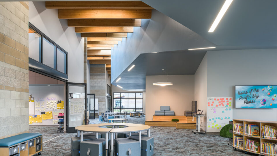 Modern school library interior with round tables, low stools, bookshelves, and a screen reading Home of the Pacific Sky Pilots!