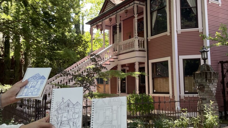 Hands hold sketchbooks with house drawings in front of a pink Victorian house shaded by large trees.