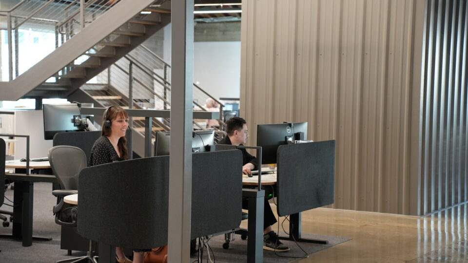 Two employees work at computers in a modern open office with partitioned desks and a staircase in the background.