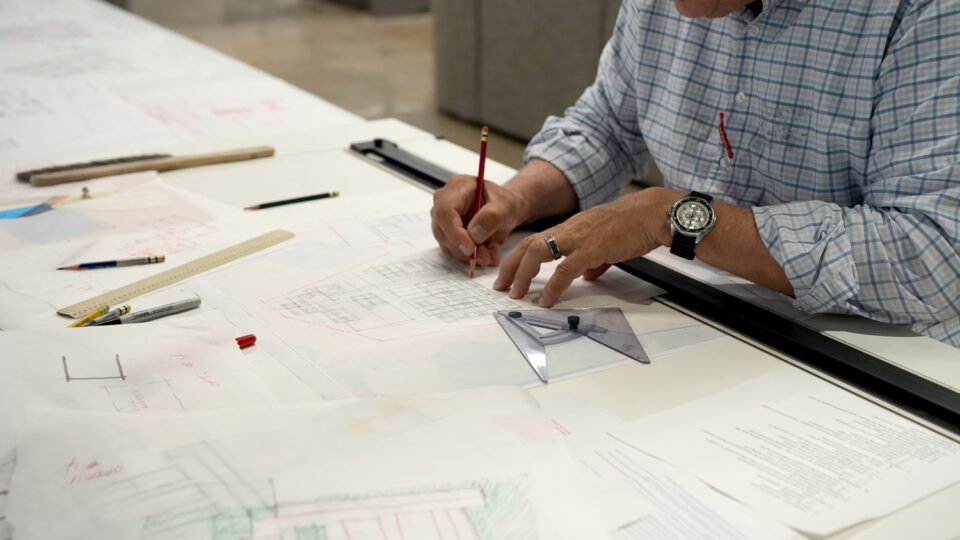 Man drawing architectural plans at a drafting table with pencils, ruler, and triangle.