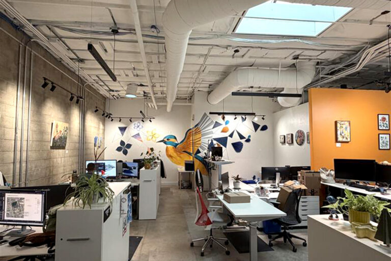 Open office with desks and computers, exposed ducts, skylights, and a large colorful bird mural on the back wall