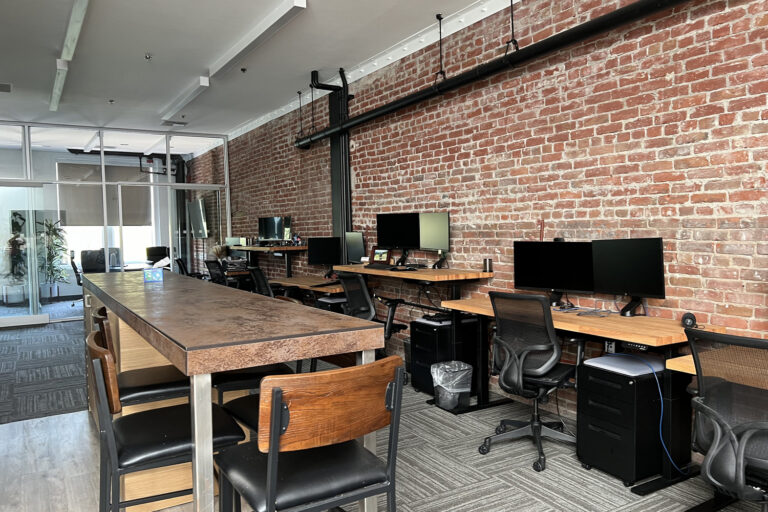 Modern office workspace with exposed brick walls, desks with computer monitors, rolling chairs, and a long high table.