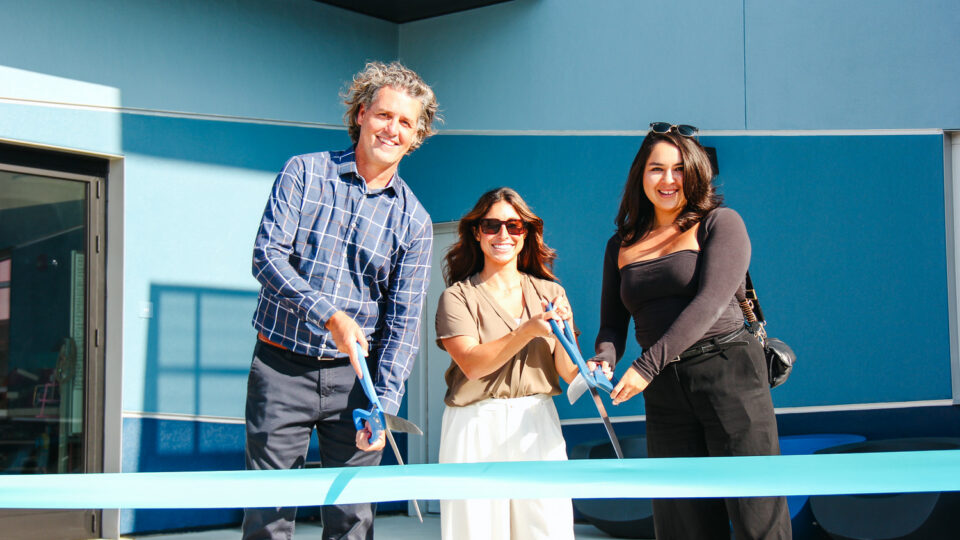 Three people smiling and cutting a blue ribbon outside a modern building opening ceremony
