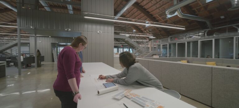 Two coworkers review plans and drawings at a long table in a spacious modern office with exposed beams.
