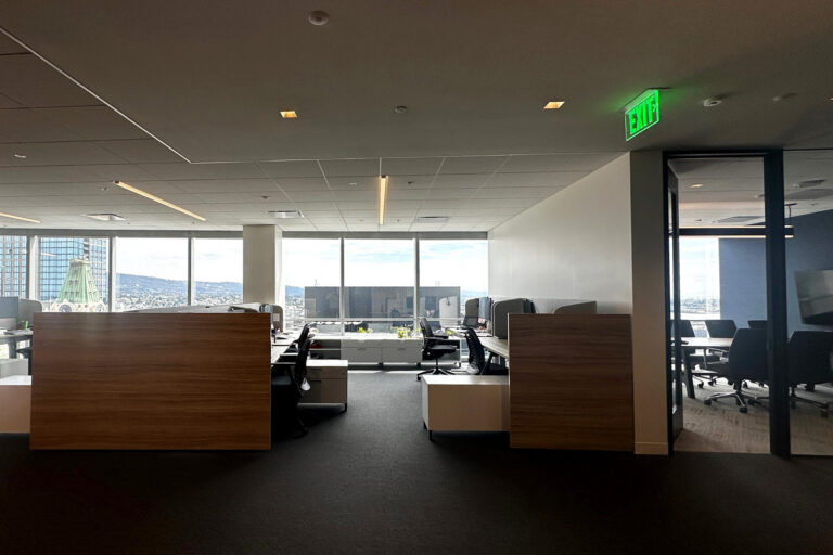 Modern office with cubicles and a glass meeting room, large windows showing a city and hillside view, and a lit EXIT sign.