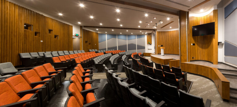 Modern auditorium with curved rows of orange and gray seats, wood-paneled walls, and a small stage with a screen