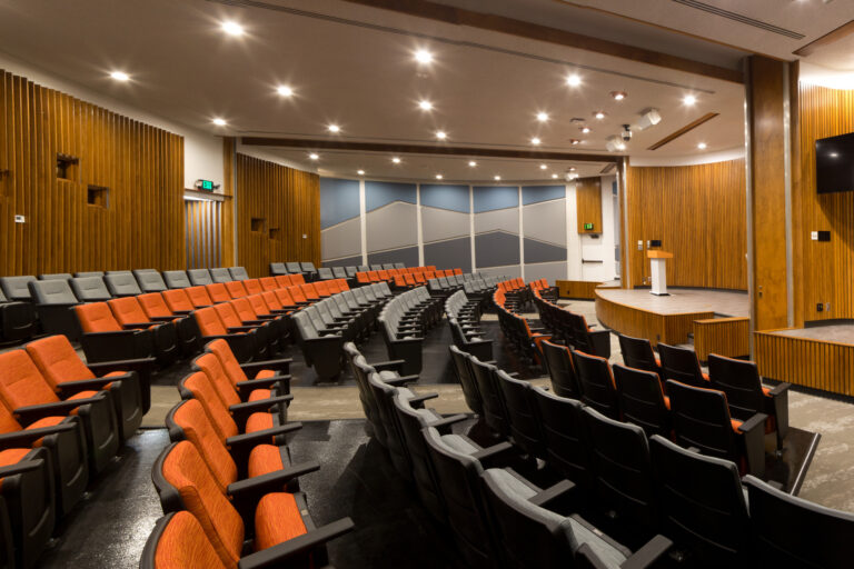 Modern auditorium with curved rows of orange and gray seats, wood-paneled walls, and a small stage with a screen