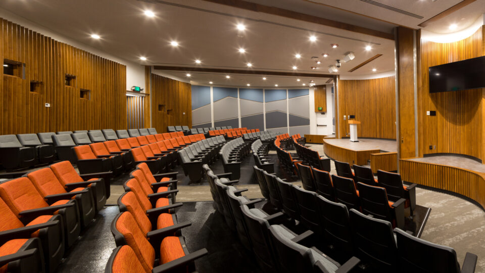 Modern auditorium with curved rows of orange and gray seats, wood-paneled walls, and a small stage with a screen