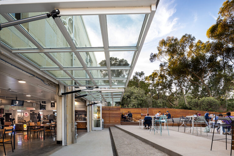 Outdoor café patio beside a building, covered by a glass awning, with scattered tables and trees in the background