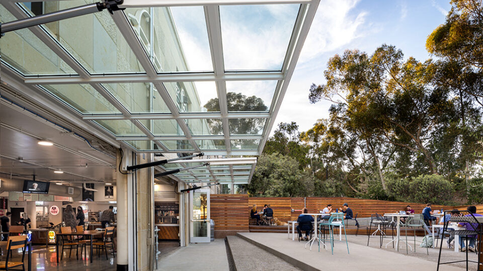 Outdoor café patio beside a building, covered by a glass awning, with scattered tables and trees in the background